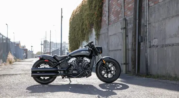 Czarny motocykl cruiser Indian Scout stoi na ulicy, otoczony miejską scenerią z ceglanym murem i zielenią.