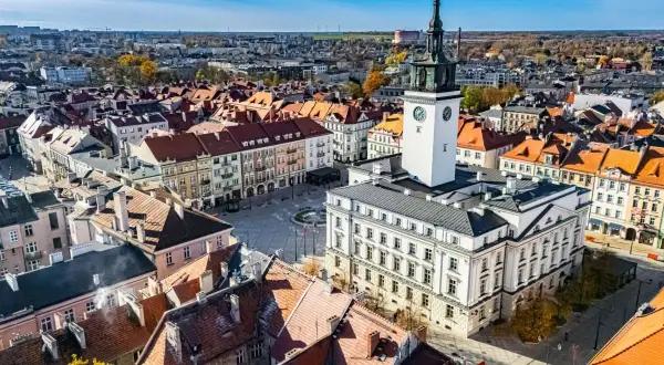 Widok z lotu ptaka na rynek z ratuszem i wieżą zegarową, otoczony kamienicami.