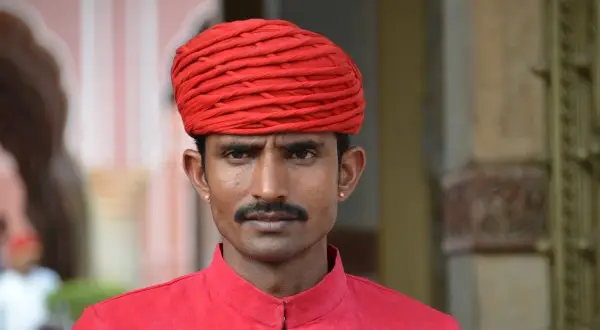 Un homme porte un turban rouge vif et une chemise assortie. Sa moustache fine souligne son regard intense.