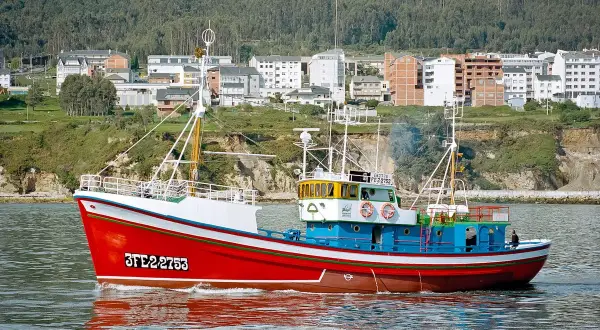 El barco museo boniteiro Reina del Carmen navega con su casco rojo y azul, reflejándose en el agua.