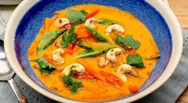 Ein cremiges rotes Thai Curry mit Cashewkernen, Zuckerschoten und Karotten in einer blauen Schüssel.