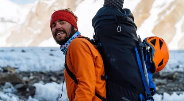 Kacper Tekieli, mit roter Mütze und orangefarbener Jacke, lächelt in die Kamera, bereit für sein nächstes Bergabenteuer.