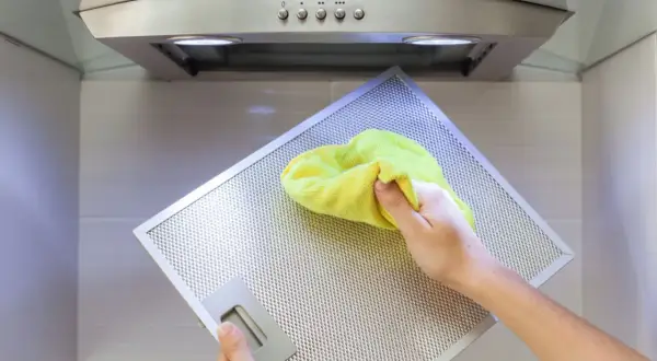 Hand reinigt mit gelbem Tuch den Metallfilter einer Dunstabzugshaube, um klebrige Rückstände zu entfernen.