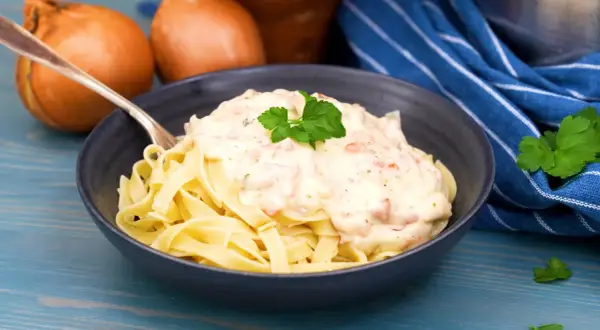 En skål med tagliatelle toppad med krämig ost och skinksås, garnerad med persilja. Två gula lökar syns i bakgrunden.
