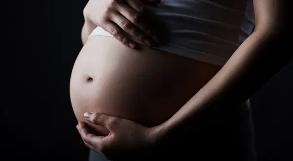 Brzuch kobiety w ciąży, z dłońmi delikatnie go obejmującymi, na ciemnym tle.
