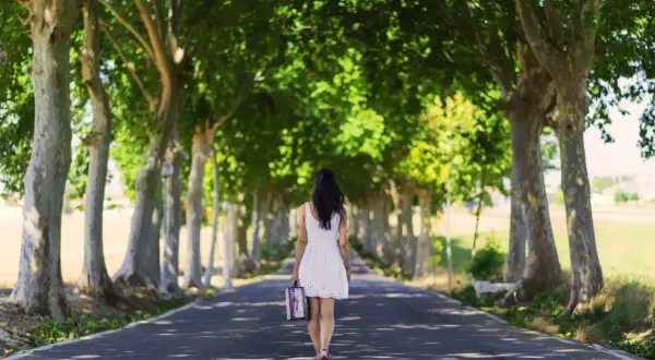 Vrouw in witte jurk loopt alleen over een laan met bomen, met een koffer in haar hand. Ze loopt weg, alsof ze weet dat ik val op mannen die mij afwijzen.