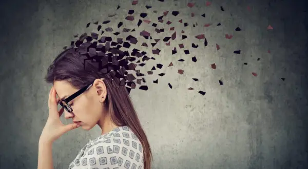 Kobieta z ręką na czole, której głowa rozpada się na fragmenty. Obraz symbolizuje **adhd z przewagą zaburzeń koncentracji uwagi**, trudności w skupieniu myśli.