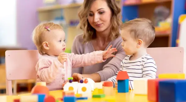 Dzieci bawią się klockami przy stole, a opiekunka patrzy z uśmiechem. Dofinansowanie do żłobka ułatwia rodzicom opiekę nad maluchami.