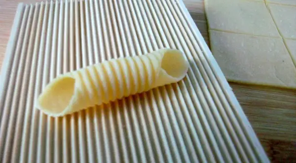 Frische Pasta selbst machen ohne Maschine: Ein geriffeltes Werkzeug formt Teig zu Röhren. Daneben liegen Teigplatten.