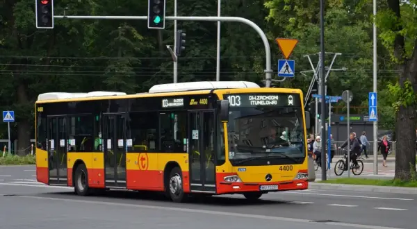Żółto-czerwony autobus linii 103 w Warszawie jedzie ulicą. W tle drzewa i sygnalizacja świetlna.