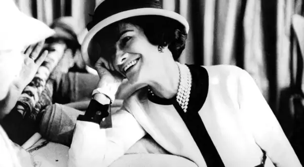 Uśmiechnięta kobieta w kapeluszu i perłach, z dłonią opartą o policzek. Ikona stylu, Coco Chanel, w swoim charakterystycznym, eleganckim stroju.
