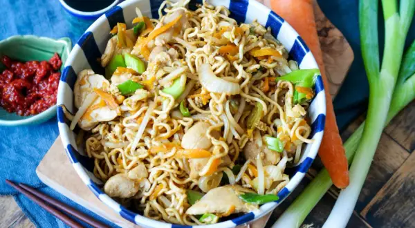 Bami Goreng mit Hühnchen, Karotten und Frühlingszwiebeln in einer Schüssel, daneben Sojasauce, Chili und frisches Gemüse.