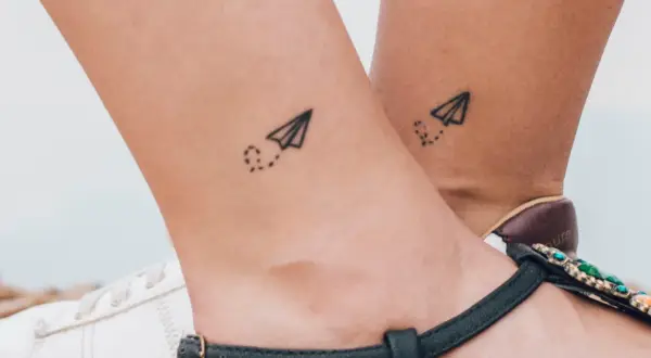 Dwa małe tatuaże samolotów z kropkowanym śladem na kostkach. Symbolizują podróże i wolność.