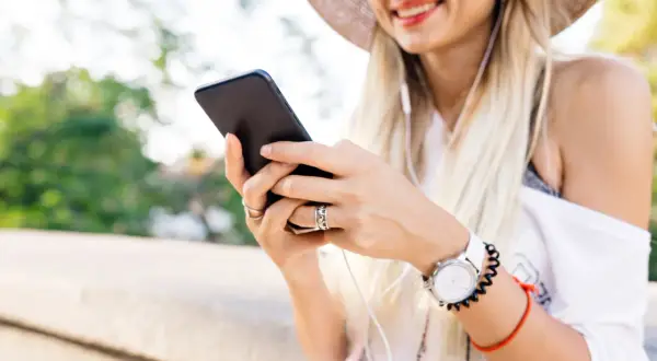 Uśmiechnięta kobieta w kapeluszu słucha muzyki przez słuchawki, trzymając telefon. Może szuka instrukcji, jak zainstalować radio w telefonie.