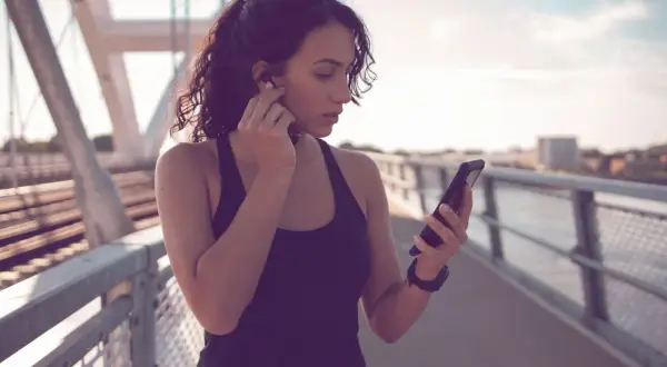 Frau auf einer Brücke, die sich Kopfhörer einsetzt und auf ihr Handy schaut. Sie ist bereit für ihre täglichen 10.000 Schritte.