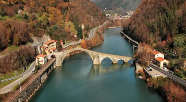 Il **Ponte del Diavolo**, con le sue arcate in pietra, attraversa un fiume circondato da colline autunnali e case.