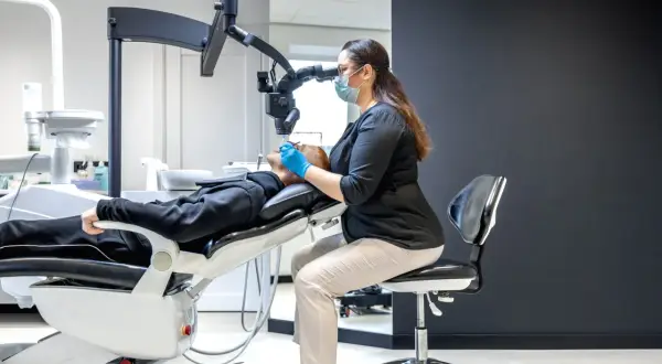 Dentystka w masce i rękawiczkach precyzyjnie pracuje pod mikroskopem, kiedy leczenie kanałowe jest wykonywane z najwyższą starannością.