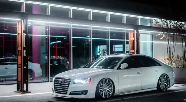 Białe Audi z obniżonym zawieszeniem **air ride** stoi przed nowoczesnym budynkiem z neonowym oświetleniem.