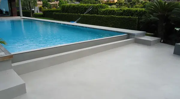 Modern villa with a bright blue pool and surrounding terraces finished with elegant piscinas de microcemento. Lush green hedges and palm trees frame the scene.