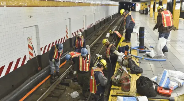 Des ouvriers en tenue de chantier, gilets de sécurité et casques, travaillent sur les voies d'une station de métro. Ils semblent engagés dans des travaux de maintenance, peut-être le prime habillage ou deshabillage de certains éléments.