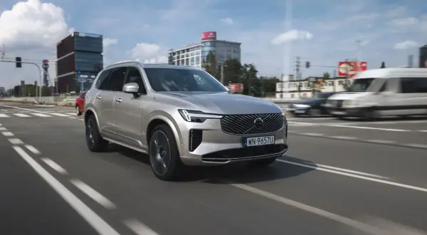 Srebrny Volvo XC90 T8 w ruchu miejskim, z nowoczesnymi budynkami w tle.
