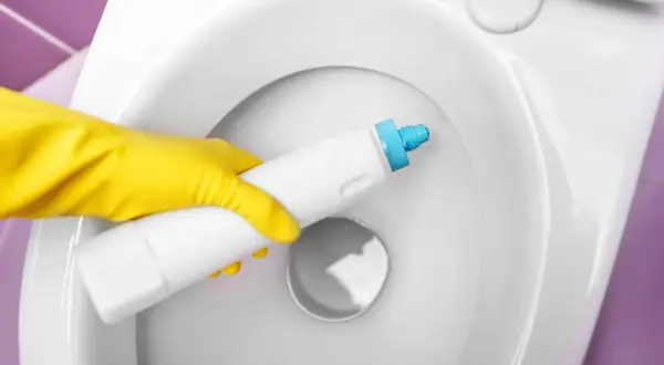 Main jaune appliquant un produit pour éliminer le calcaire dans une toilette blanche.
