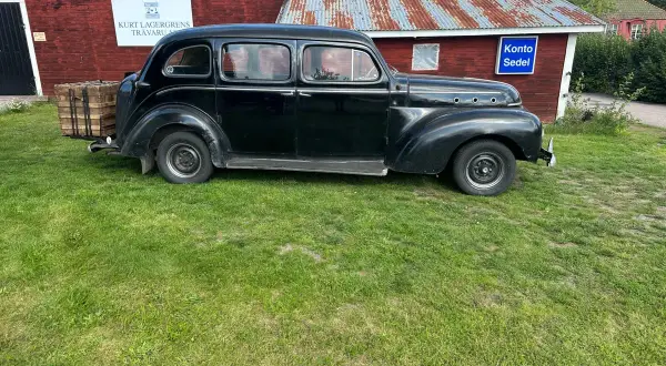 Svart Volvo PV 831 parkerad på gräs framför en röd byggnad med rostigt plåttak. En träkorg sitter på bakre stötfångaren.