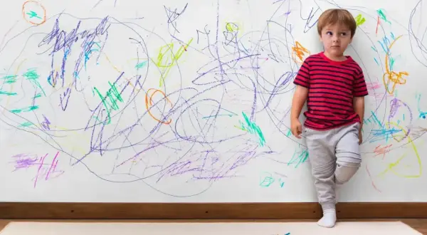 Chłopiec stoi przy ścianie pokrytej kolorowymi bazgrołami. Czy to oznaka, jak rozpoznać ADHD u dziecka?