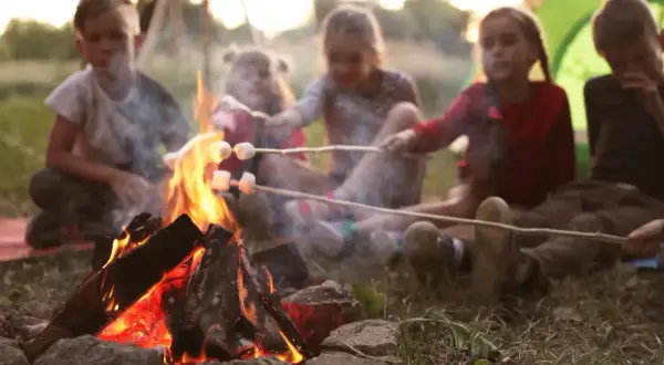 Kinder rösten Marshmallows über einem Lagerfeuer. Ein Zelt ist im Hintergrund zu sehen.