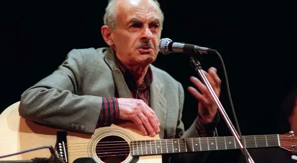 Starszy mężczyzna z gitarą i mikrofonem, śpiewający piosenkę o wojnie.