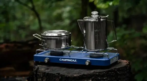 Niebieska kuchenka turystyczna Campingaz z dwoma palnikami, na której stoją metalowy garnek i kawiarka. Idealna na biwak.