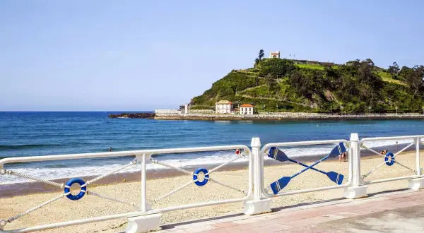 La **playa de Santa Marina** con su arena dorada, el mar azul y una colina verde coronada por un edificio blanco.