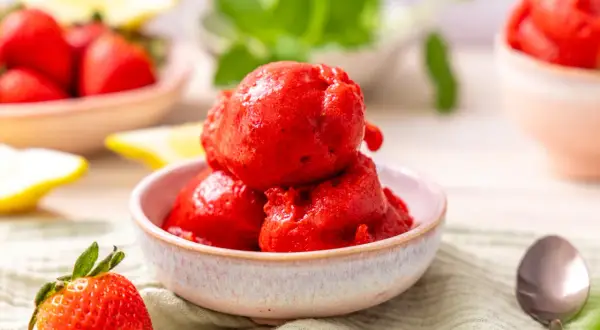 Schüssel mit leuchtend rotem Erdbeer-Sorbet, perfekt zum Sorbet selber machen. Frische Erdbeeren und Zitronenspalten im Hintergrund.