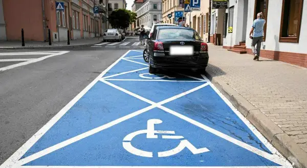 Niebieskie pole z białym symbolem wózka inwalidzkiego to **miejsce dla niepełnosprawnych**. Zaparkowany czarny samochód zajmuje ten specjalny parking.
