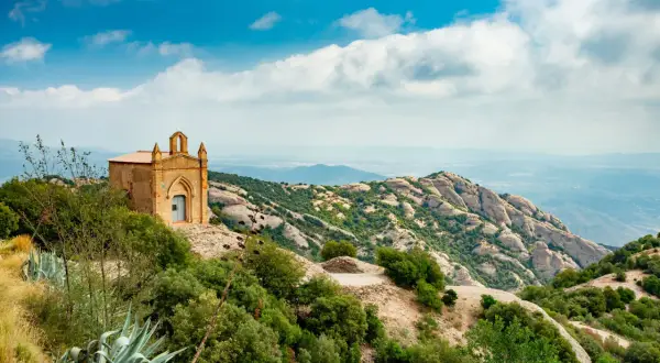 Pequeña ermita en la cima de una montaña, ideal para un **montserrat hike barcelona**. Paisaje rocoso y verde con cielo azul y nubes.