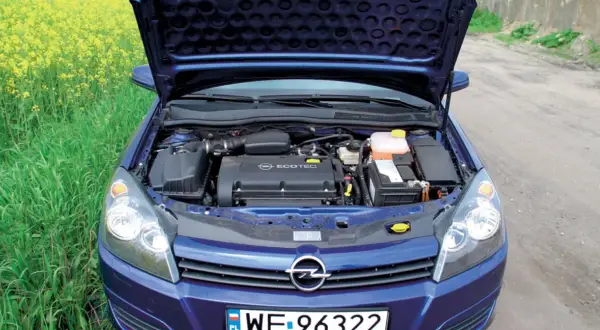 Silnik Ecotec w niebieskim Opel Astra H 1.6 benzyna, z otwartą maską, na tle pola rzepaku.