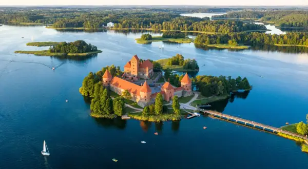 Zamek Troki, malowniczo położony na wyspie pośród jeziora, otoczony zielenią i wodą, z mostem prowadzącym do jego bram.