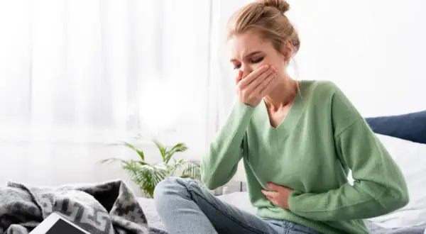 Mujer rubia con moño sentada en la cama, con la mano en la boca y otra en el abdomen, sufriendo nauseas antes de la regla.