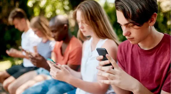 Grupa nastolatków siedzi na ławce, każdy pochłonięty smartfonem. Warto zadbać o ich bezpieczeństwo online, np. poprzez kontrolę rodzicielską na Androidzie.