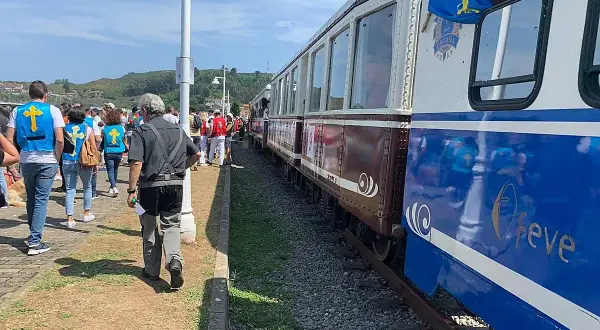 Gente con chalecos amarillos espera el tren descenso del Sella. El tren tiene vagones marrones y azules con detalles dorados.
