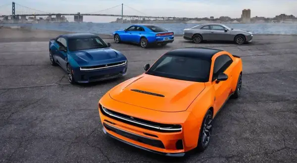 Pomarańczowy i niebieski Dodge Charger Hellcat z 2024 roku, obok szarego coupe, na tle mostu i miasta.