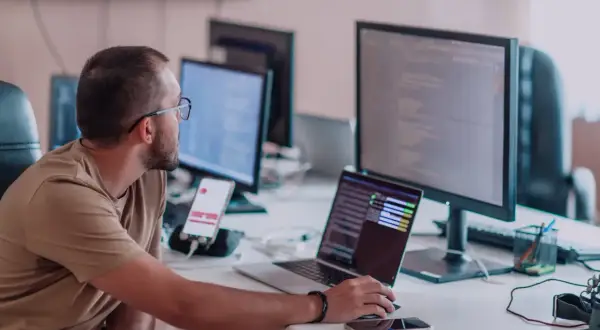 Programista aplikacji w okularach pracuje przy biurku z kilkoma monitorami i laptopem.