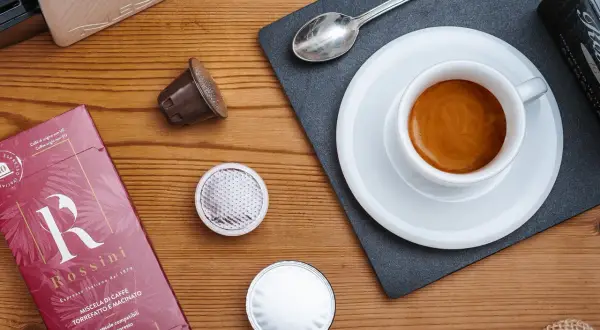 Una tazzina di espresso fumante, capsule e confezione Rossini: un invito a gustare una nota varietà di caffè.