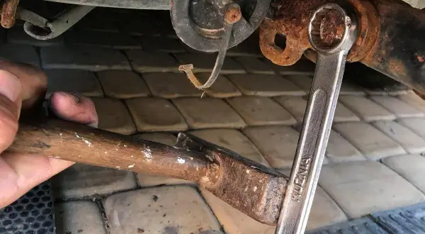 Ręka próbuje odkręcić zapieczoną śrubę kluczem, używając młotka do pomocy.