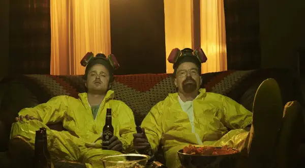 Die Besetzung von Breaking Bad, Walter und Jesse, entspannt in ihren Schutzanzügen mit Bier und Snacks auf dem Sofa.