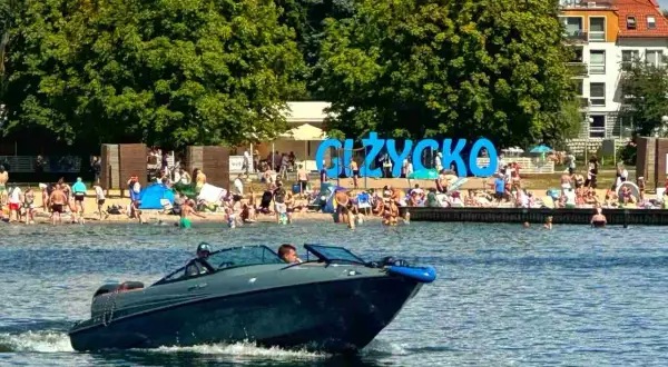 Łódź motorowa na jeziorze, plaża pełna ludzi i wielki napis GIŻYCKO. Idealne miejsce, gdzie na Mazury!