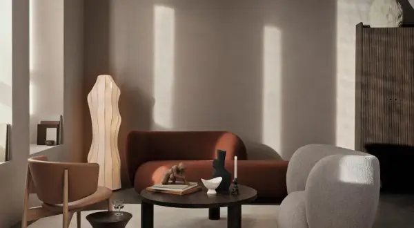 Ein stilvolles Wohnzimmer mit einer rostbraunen Couch. Ein beiger Bouclé-Sessel und ein Holzstuhl ergänzen die Sitzgelegenheiten. Ein runder Couchtisch mit Deko-Objekten steht im Zentrum.