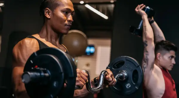 Zwei Männer trainieren im Fitnessstudio. Der vordere Mann hebt eine Hantel, der hintere Mann stemmt eine Kurzhantel. Verschiedene Kraft Arten werden hier gezeigt.