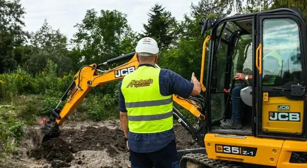 Instruktor z Akademii Operatora pokazuje, ile kosztuje godzina pracy koparki JCB. Operator w kabinie.