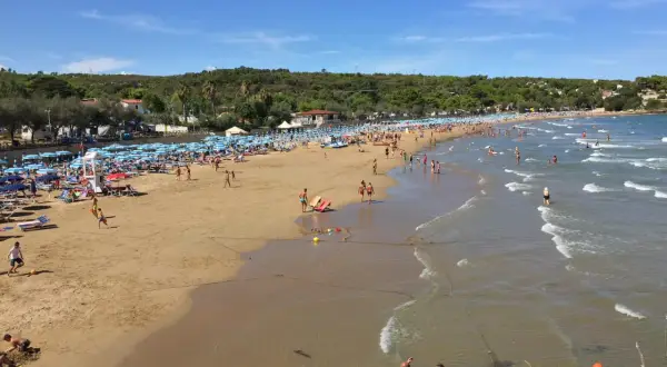 Una giornata di sole sulla **spiaggia di Bescile**, con ombrelloni colorati, bagnanti che giocano e si rilassano, e il mare che invita a un tuffo.